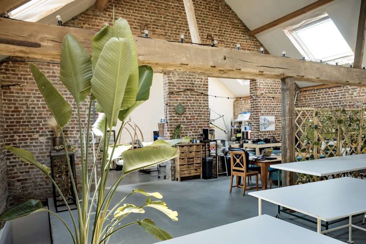 De studio: een oude schuur met houten spanten, een dakraam en bakstenen muren, met witte werktafels onder de balken.